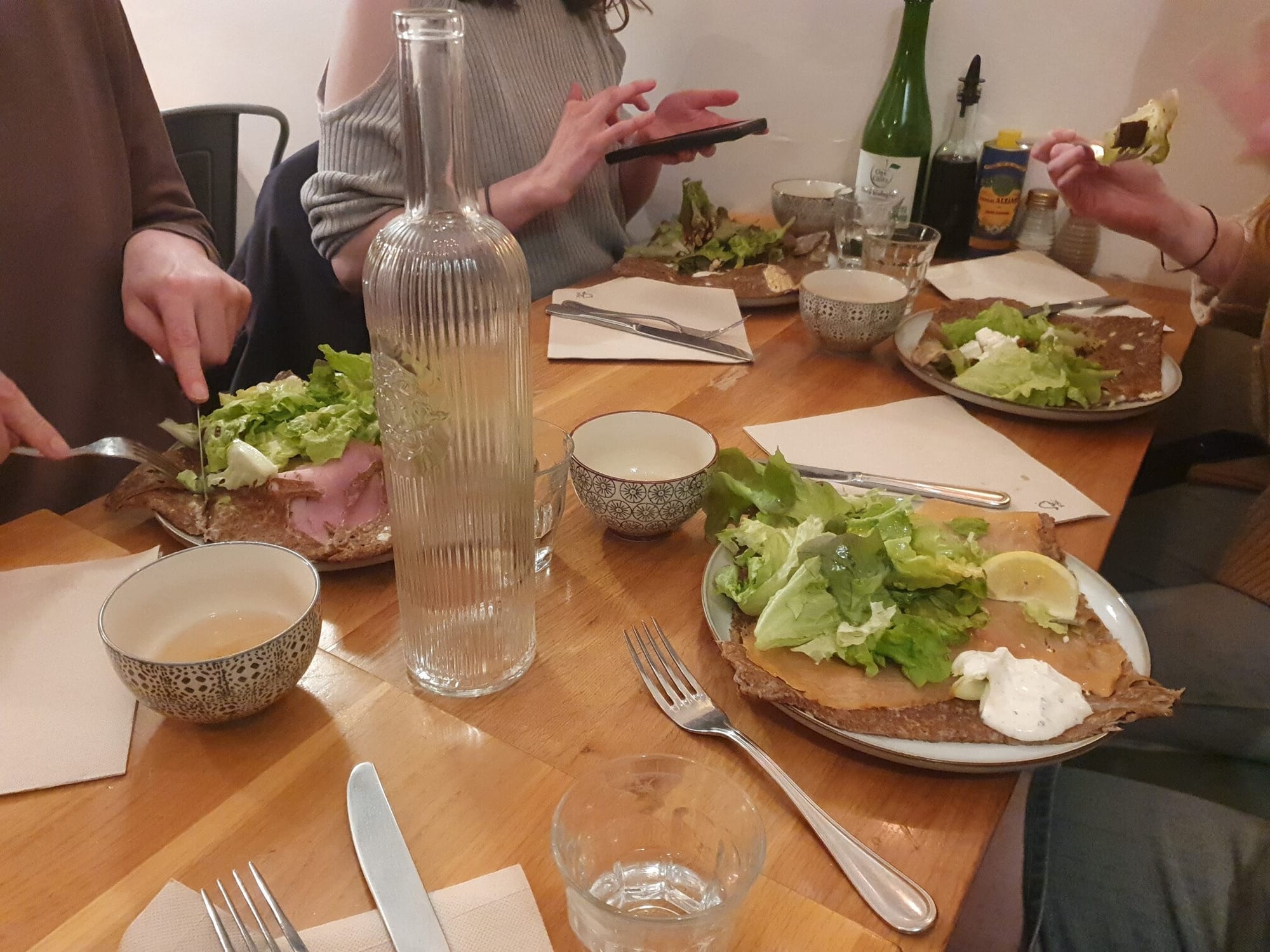 A wooden table, filled with cutlery and plates with giant dark crepes filled with salad