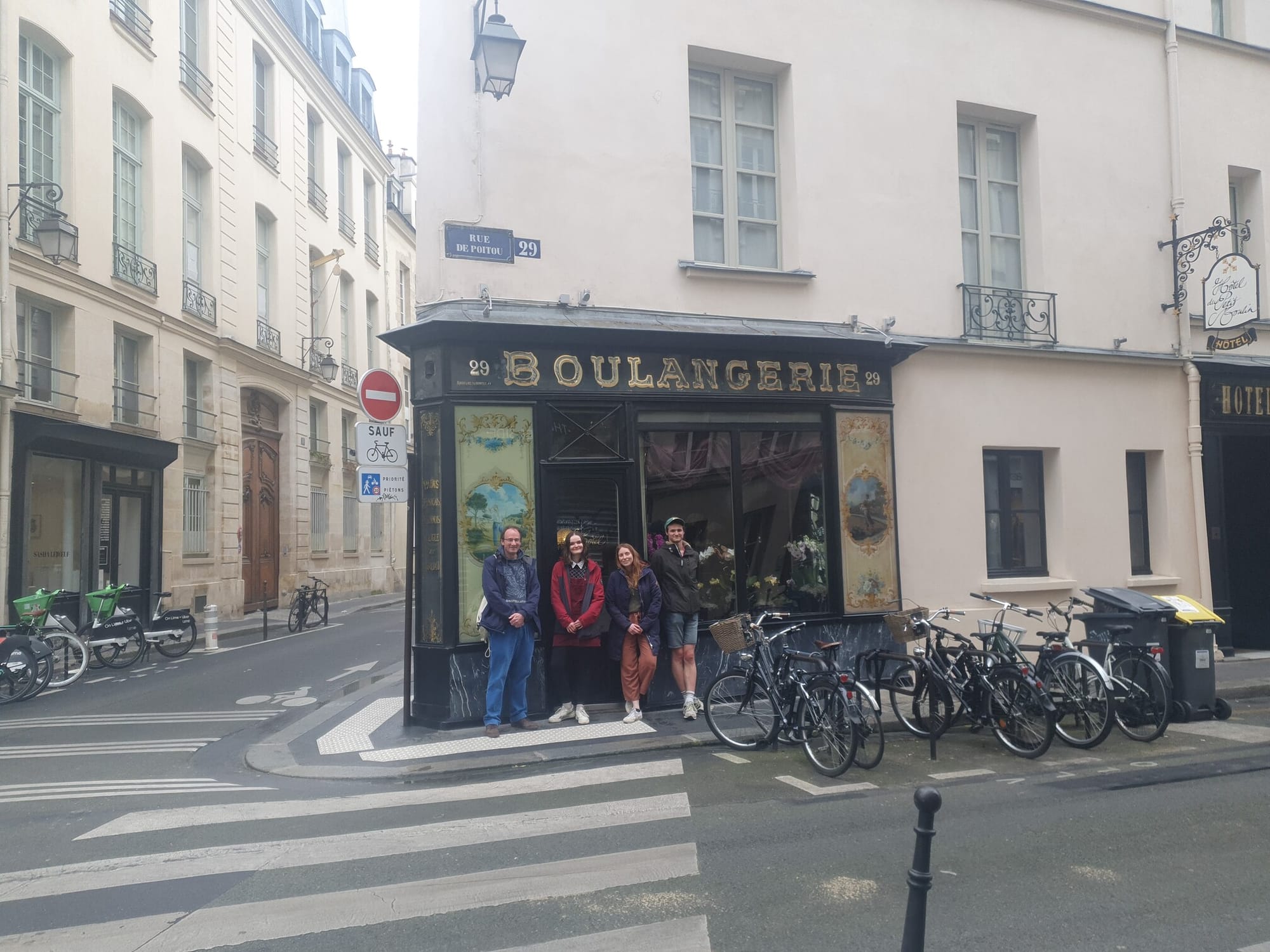 4 people and a lot of bikes, outside an old painted black shop, with 'Boulangerie' written in gold lettering