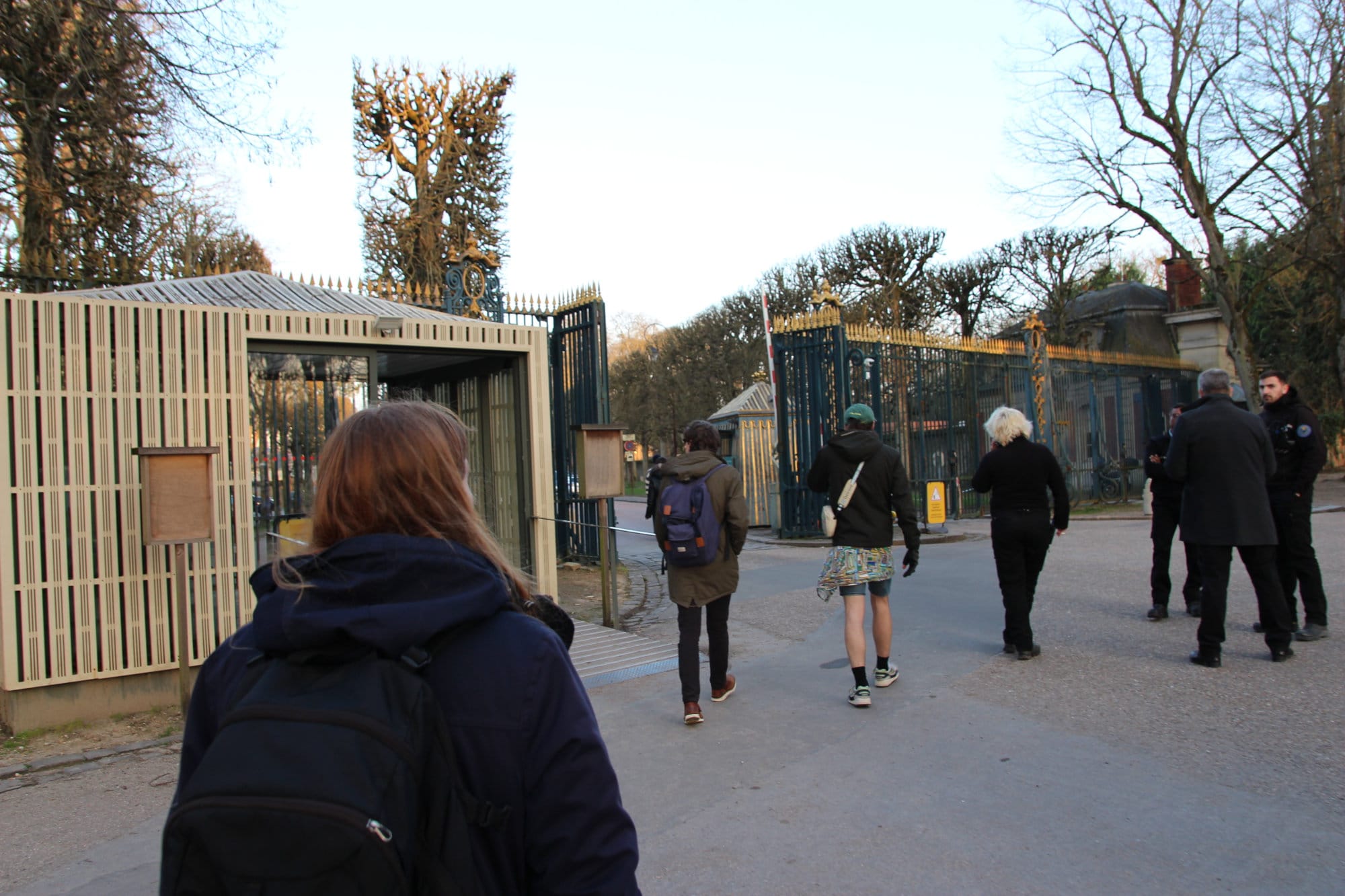 Backs of people walking through a gold topped fence and an open gate, with sky darkening and police in tow!