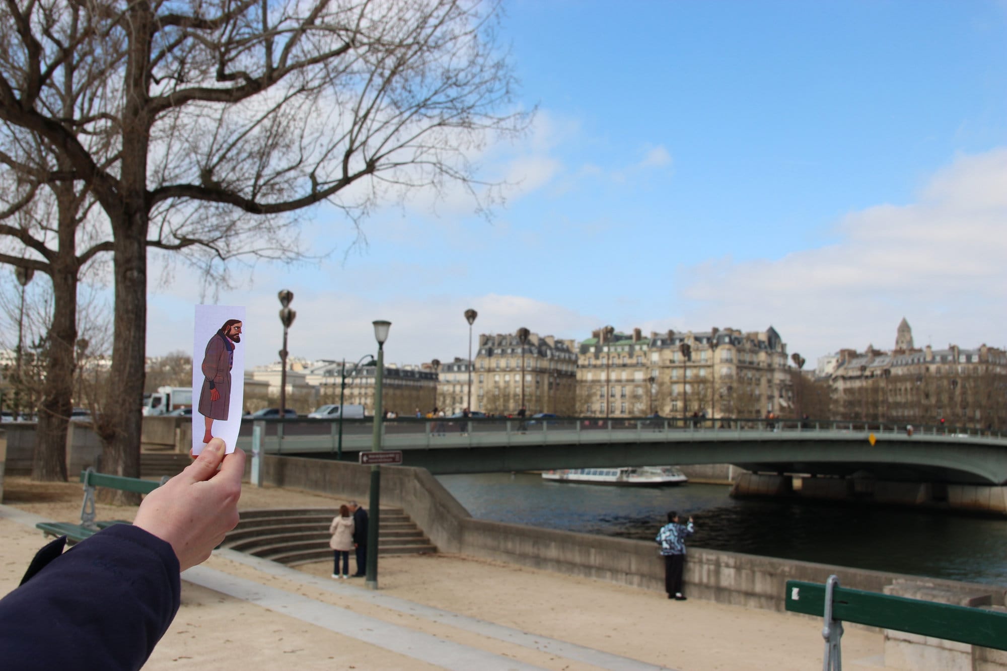 Paper cutout of a man in a long brown coat, 'walking' across a bridge in Paris